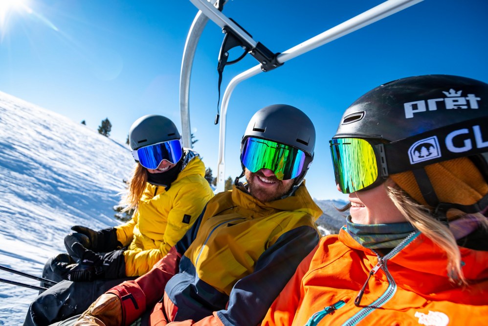 3 adults in ski lift wearing helmets and goggles